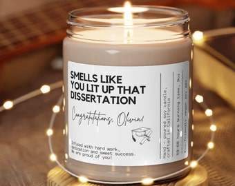 May include: A brown glass candle with a white label that reads "Smells like you lit up that dissertation. Congratulations, Olivia!" The label also includes a small illustration of a graduation cap and diploma. The candle is infused with hard work, dedication, and sweet success. It is a 9 oz. hand-poured soy candle with a 50-60 hour burn time.