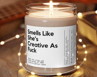 May include: A brown glass candle with a white label that reads "Smells Like She's Creative As Fuck". The label also includes the text "Snell the crisp and fresh aroma of 100% creative genius vibes".