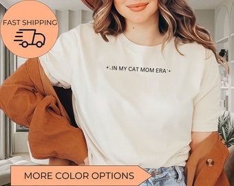 May include: White t-shirt with the text "*IN MY CAT MOM ERA*" printed on the front. The shirt is being worn by a person with long brown hair and a brown jacket.