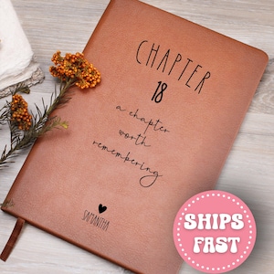 May include: A brown leather journal with the text "CHAPTER 18" and "a chapter worth remembering" in black script. The journal also has the name "SAMANTHA" and a heart symbol. A sprig of orange flowers rests on the journal.
