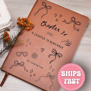 May include: A brown leather journal with the text "Chapter 14: A Chapter to Remember" and the name "By: Samantha" in black. The journal is decorated with black line drawings of bows, hearts, stars, sunglasses, and a camera. A brown ribbon bookmark is visible.