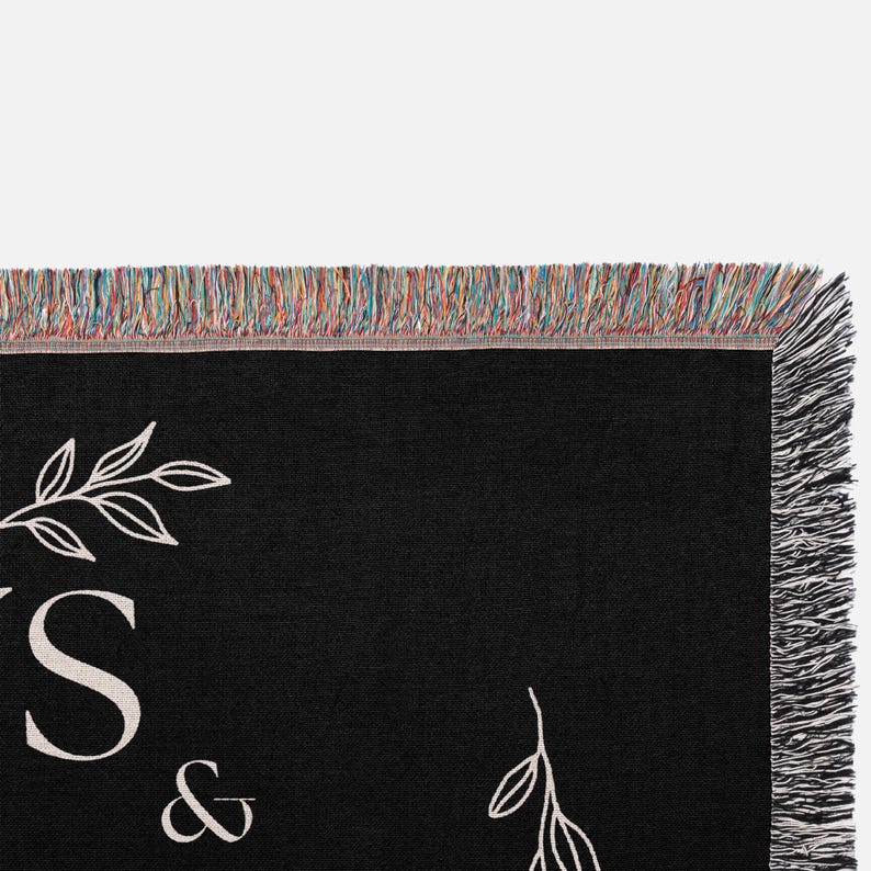 May include: Black woven throw blanket with white floral designs and the text "&" in the centre. The blanket has a fringe border.