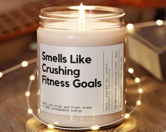 May include: A white soy candle with a black label that reads "Smells Like Crushing Fitness Goals". The label also includes the text "Hand-poured soy candle, crafted in California. 50-60 hours burning time, 9oz. Eco-friendly vegan soy wax".