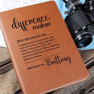 May include: A brown leather journal with the words "difference maker" and a definition of the word. The name "Brittany" is written below the definition. The journal is lying on a brown surface.