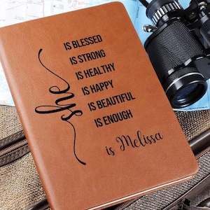 May include: A brown leather journal with the text "She is Blessed, Strong, Healthy, Happy, Beautiful, Enough, is Melissa" in black. The journal is next to a pair of binoculars and a map.