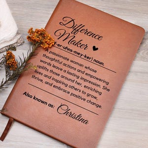 May include: A brown leather journal with the words "Difference Maker" and a definition printed on the cover. The journal includes the name "Christina" and a ribbon bookmark. A sprig of orange flowers rests on the journal.