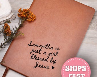 Puede incluir: Un diario de cuero marrón con la inscripción "Samantha is just a girl blessed by Jesus." Un círculo rosa con las palabras "SHIPS FAST" es visible. El diario tiene una cinta de marcador marrón y está decorado con una ramita de flores naranjas.