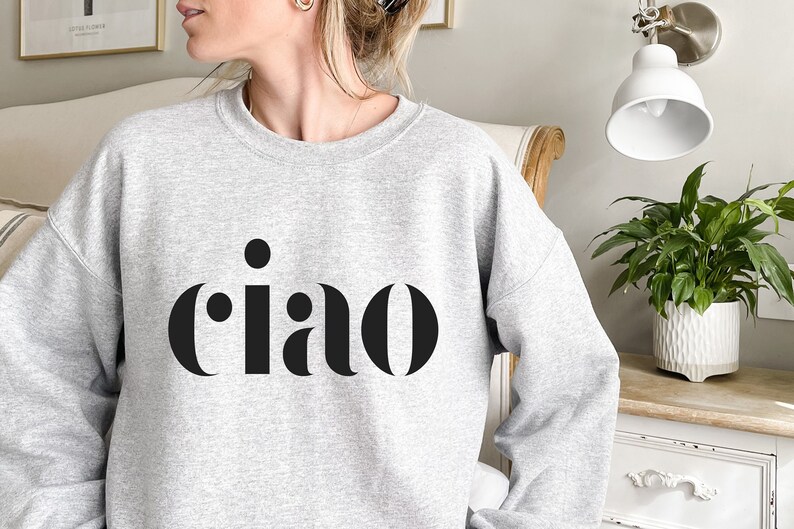 May include: A pale grey crew neck sweatshirt featuring the word "ciao" in bold, black lettering. The Italian greeting is centrally placed on the chest. The image is well-lit, and the sweatshirt appears to be made of a soft material, suitable for casual wear.