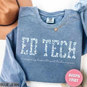 May include: A blue jean colored sweatshirt with the words "ED TECH" in large, floral-patterned letters. Below the text is the phrase "emergency department technician" in a cursive font. A gold necklace with a pendant is draped on the sweatshirt.