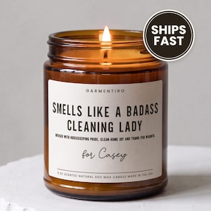 May include: A 9 oz scented natural soy wax candle in an amber glass jar with a lit wick. The white label features the text GARMENTIRO, SMELLS LIKE A BADASS CLEANING LADY, INFUSED WITH HOUSEKEEPING PRIDE, CLEAN-HOME JOY AND THANK-YOU WARMTH, for Casey.