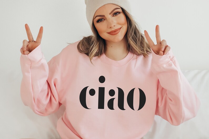 May include: A pale pink sweatshirt featuring the word "ciao" in black, bold lettering. The person in the image is wearing a light beige beanie and making a peace sign with their fingers.