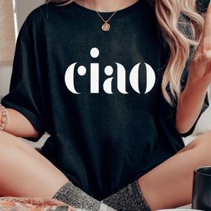 May include: Black t-shirt with the word "ciao" in large, white, bold letters. The shirt is worn by a person sitting cross-legged. The person is wearing a necklace and bracelets. The person is also giving a peace sign.