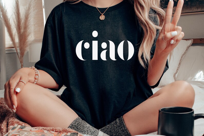 May include: Black t-shirt with the word "ciao" in large, white, bold letters. The shirt is worn by a person sitting cross-legged. The person is wearing a necklace and bracelets. The person is also giving a peace sign.