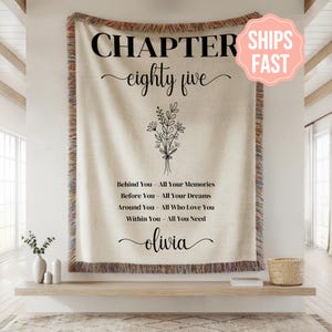 Puede incluir: Una manta beige con texto negro que dice "CHAPTER eighty five" y una ilustración floral. Debajo de la ilustración está el texto "Behind You - All Your Memories, Before You - All Your Dreams, Around You - All Who Love You, Within You - All You Need, olivia."