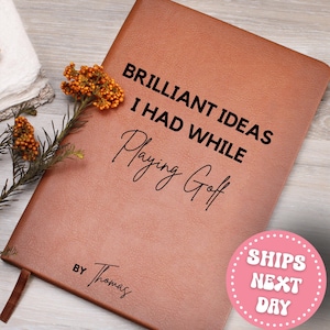 May include: A brown leather journal with the text "BRILLIANT IDEAS I HAD WHILE Playing Golf" in black. The journal has a ribbon bookmark and the name "Thomas" at the bottom. A sprig of orange flowers and greenery is on the left.