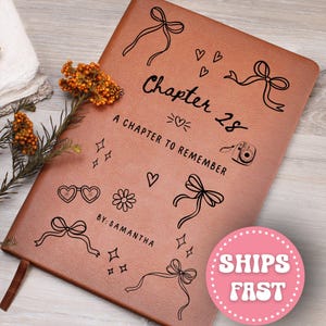May include: A brown leather journal with black line art of bows, hearts, stars, and a camera. The text "Chapter 28, A Chapter to Remember, By: Samantha" is printed on the cover. A pink circle with "Ships Fast" is in the bottom right corner.