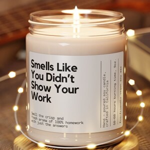 May include: A white soy candle with a black label that reads "Smells Like You Didn't Show Your Work". The candle is hand-poured and crafted in California. It has a 50-60 hour burn time and is made with eco-friendly vegan soy wax.
