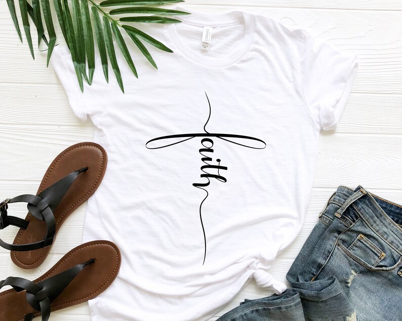 May include: White t-shirt with a black cross and the word "faith" written vertically on it. The shirt is laid flat on a white wooden surface with a pair of brown sandals and blue jeans.