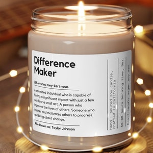 May include: A brown glass candle jar with a white label that reads "Difference Maker (dif-er-uhns-mey-ker) noun. A committed individual who is capable of having a significant impact with just a few words or a small act. A person who improves the lives of others. Someone who inspires and motivates others to progress and bring about change. Also known as: Taylor Johnson". The label also says "Hand poured soy candle, crafted in California. 50-60 hours burning time, 9oz. Eco-friendly vegan soy wax".