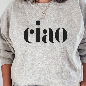 May include: A pale grey sweatshirt featuring the word "ciao" in large, black, sans-serif letters. The word is centered on the chest. The sweatshirt has long sleeves and a round neck.