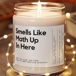 May include: A lit soy wax candle in a clear glass jar with a white label that reads "Smells Like Math Up In Here." The candle is surrounded by string lights and sits on a wooden base. The label also includes the text "Smell the crisp and fresh aroma of 100% numerical precision."