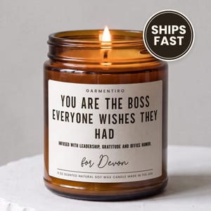 May include: A 9 oz amber glass jar candle with a lit wick. The white label features the text: GARMENTIRO, YOU ARE THE BOSS EVERYONE WISHES THEY HAD, INFUSED WITH LEADERSHIP, GRATITUDE AND OFFICE HUMOR, for Devon. The candle is made of natural soy wax.