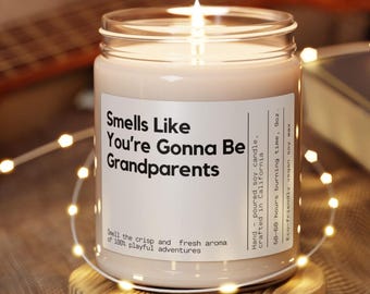 May include: A clear glass candle jar with a white label that reads "Smells Like You're Gonna Be Grandparents". The candle is a light beige color and is lit. The label also includes the text "Hand-poured soy candle, crafted in California, 50-60 hours burning time, 9oz. Eco-friendly vegan soy wax."