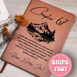May include: A brown leather journal with the words "Chapter 67" and "Happy Birthday, John!" in black script. The journal features a mountain and lake design. The text reads, "Celebrate everything you've become..."