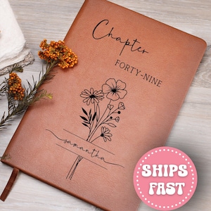May include: A brown leather journal with the words "Chapter FORTY-NINE" and a floral design. The name "Samantha" is written below the flowers. A brown ribbon bookmark is visible.