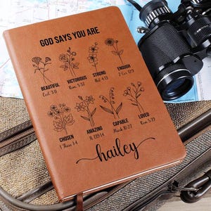 May include: A brown leather journal with the words "God Says You Are" and a list of positive affirmations with bible verses. The journal is personalized with the name "hailey".