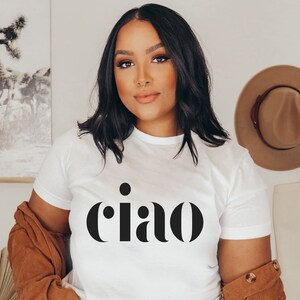 May include: White t-shirt with the word "ciao" in black, a brown jacket, and a tan hat hanging on the wall. A tote bag hangs next to the hat. A framed picture of a desert landscape is visible in the background.