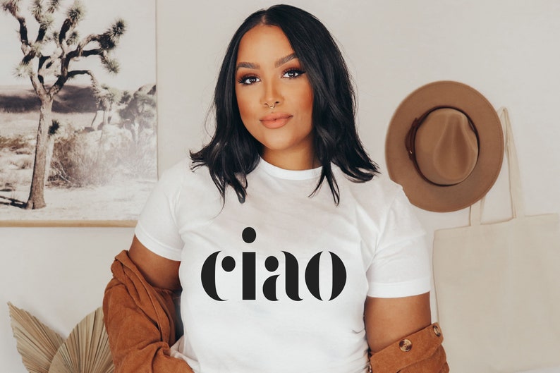 May include: White t-shirt with the word "ciao" in black, a brown jacket, and a tan hat hanging on the wall. A tote bag hangs next to the hat. A framed picture of a desert landscape is visible in the background.