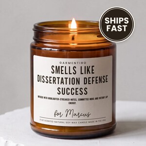 May include: A 9 oz amber glass jar candle with a lit wick. The white label reads: GARMENTIRO SMELLS LIKE DISSERTATION DEFENSE SUCCESS. INFUSED WITH HIGHLIGHTER-STREAKED NOTES, COMMITTEE NODS AND VICTORY LAP ENERGY. for Marcus. 9 OZ SCENTED NATURAL SOY WAX CANDLE.