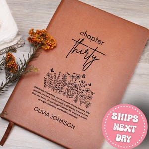 May include: A brown leather journal with the words "chapter thirty" in black script. Below the text is a floral design and the name "OLIVIA JOHNSON". The journal is open, revealing a blank page. A sprig of orange flowers rests on the side.