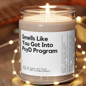 May include: A brown glass candle with a white label that reads "Smells Like You Got Into PsyD Program". The label also includes the text "Hand-poured soy candle, crafted in California, 50-60 hours burning time, 9oz. Eco-friendly vegan soy wax".