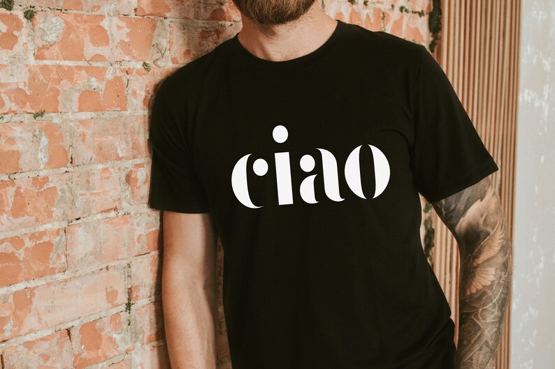 May include: Black t-shirt featuring the word "ciao" in a white, decorative font. The t-shirt has a crew neck design. The person wearing the shirt is leaning against a brick wall.