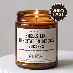 May include: A lit 9 oz scented natural soy wax candle in an amber glass jar with a white label reading "GARMENTIRO SMELLS LIKE DISSERTATION DEFENSE SUCCESS INFUSED WITH MIDNIGHT RESEARCH, BOLD IDEAS AND COMMITTEE APPLAUSE. for Rosa 9 OZ SCENTED NATURAL SOY WAX CANDLE MADE IN THE USA."