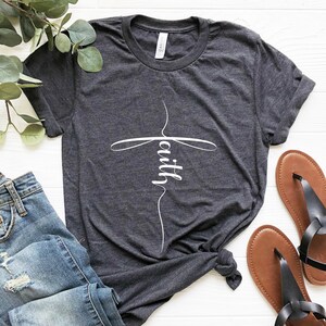 May include: A dark heather grey t-shirt with a white cross design and the word "faith" written inside the cross.