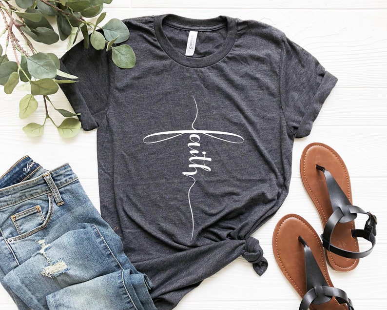 May include: A dark heather grey t-shirt with a white cross design and the word "faith" written inside the cross.