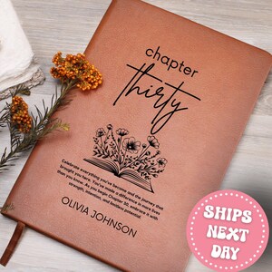 May include: A brown leather journal with the words "chapter thirty" in black script. Below the text is a floral design above the text "OLIVIA JOHNSON". The journal has a ribbon bookmark and a quote about celebrating life's journey.
