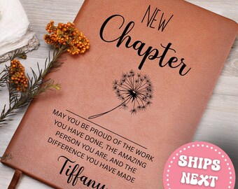 May include: A brown leather journal with the words "NEW Chapter" in elegant script. Below, a dandelion illustration and the text "MAY YOU BE PROUD OF THE WORK YOU HAVE DONE...Tiffany". A sprig of orange flowers rests on the side.