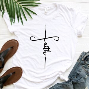 K&ouml;nnte beinhalten: Wei&szlig;es T-Shirt mit einem schwarzen Kreuz und dem Wort "faith" vertikal in der Mitte des Kreuzes.