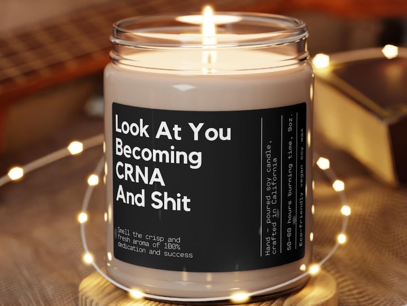 May include: A brown soy candle with a black label that reads "Look At You Becoming CRNA And Shit". The label also includes information about the candle, such as the fact that it is hand-poured and made in California. The candle is sitting on a wooden surface with string lights around it.