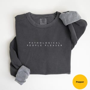 May include: A dark gray sweatshirt with the text "Pathological People Pleaser" printed in white on the front.