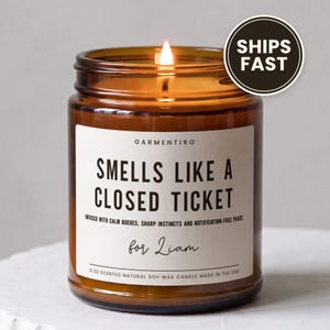 May include: A lit candle in an amber glass jar with a white label reading "GARMENTIRO SMELLS LIKE A CLOSED TICKET INFUSED WITH CALM QUEUES, SHARP INSTINCTS AND NOTIFICATION-FREE PEACE for Liam 9 OZ SCENTED NATURAL SOY WAX CANDLE MADE IN THE USA."
