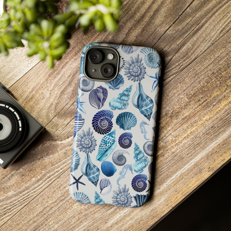 May include: A white phone case with a blue and white seashell pattern. The case has a camera lens cutout and a white background.