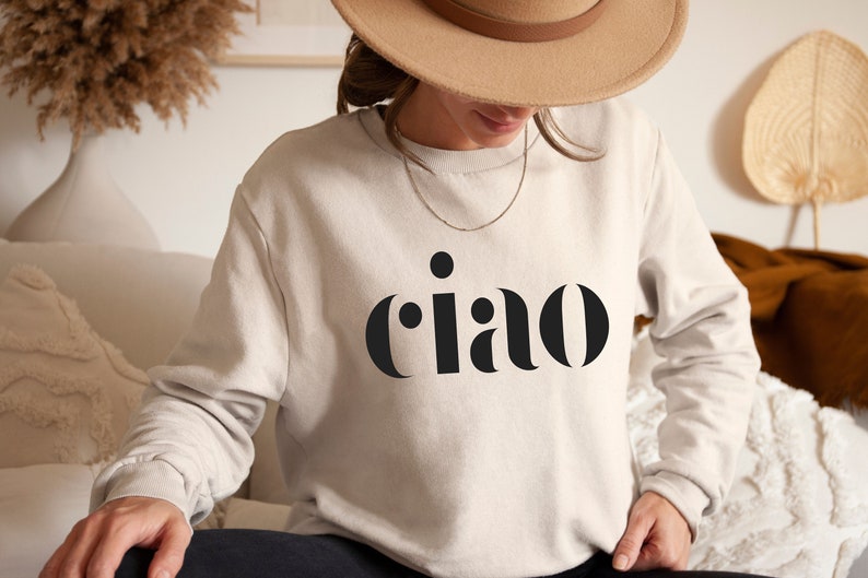 May include: A beige sweatshirt with the word "ciao" in bold, black letters. The long-sleeved sweatshirt is made of a soft material. The person is wearing a hat and a necklace. The word "ciao" is an Italian greeting.