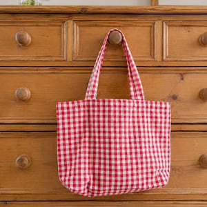 May include: A rectangular fabric tote bag featuring a classic red and white gingham checked pattern. The bag includes two matching fabric handles attached to the top edge, allowing it to be hung from a wooden knob on a piece of furniture.