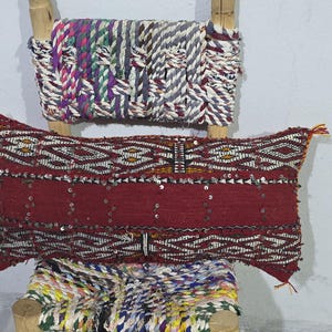 May include: A decorative, rectangular red pillow with geometric patterns and silver sequins. The pillow is adorned with tassels on the corners. It rests on a wooden structure with colorful woven textiles.
