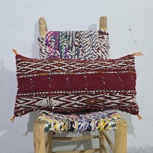 Peut inclure: Un coussin rectangulaire bordeaux avec un motif géométrique blanc et jaune, orné de paillettes. Il est posé sur une chaise en bois avec une assise et un dossier tissés. La chaise a un dossier tissé rayé coloré.